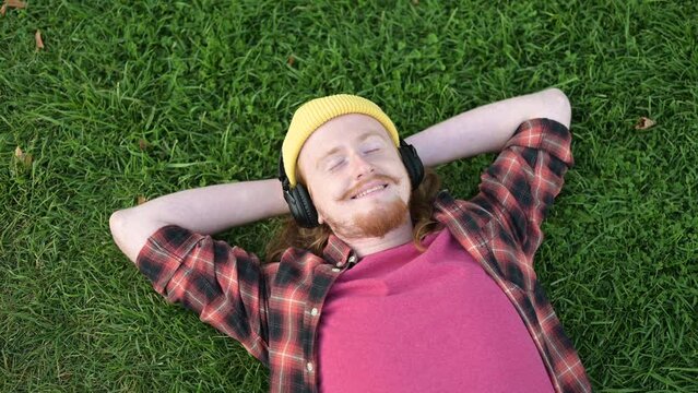 A red-haired guy in a yellow hat lies down on the grass and enjoys a summer day, listening to music on headphones.
