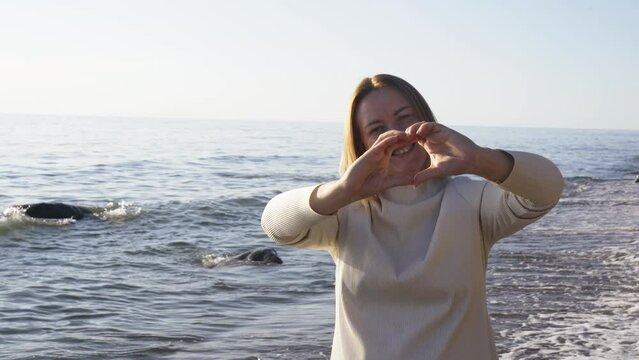 A 40-year-old woman stands on the seashore showing a heart gesture with her hands. Gestures and symbols.