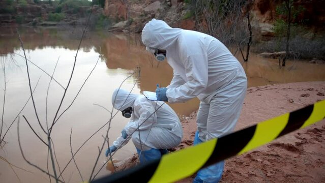 Two scientists wearing PPE, one of them sat down and scooped up water from a contaminated water source and brought it up to examine.