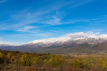 Autumn landscape showcasing trees adorned with golden foliage, with a distant high mountain peak covered in snow. Above, passing clouds in a blue sky on a sunny day in nature