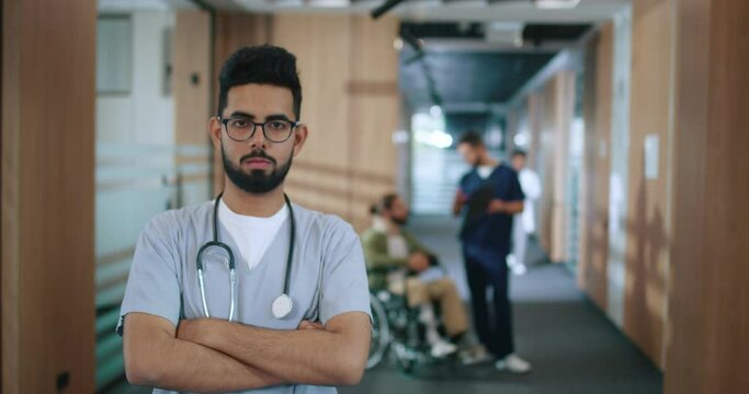 Portrait of highly qualified and professional doctor standing in hospital corridor with stethoscope and glasses. Male physiotherapist looking at camera with crossed arms.