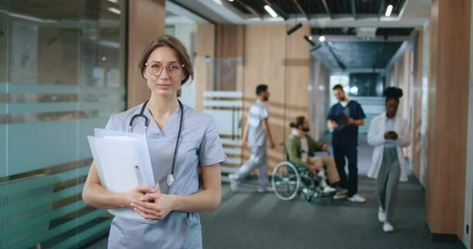Portrait of qualified cute young woman working in modern hospital wearing uniform looking at camera. Beautiful female doctor with stethoscope holding documents and standing at corridor.
