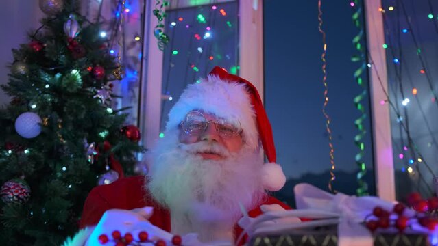 Angry bad Santa shows obscene gestures with his hands fingers sitting next to pile of New Year's gifts and a Christmas tree holiday background. 