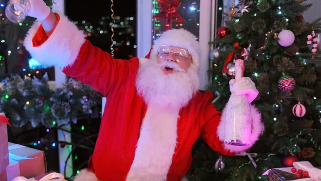 Funny fat white-haired Santa dancing with champagne and glasses in tattooed hands, celebrating Christmas in room decorated Christmas tree and beautiful light garlands.