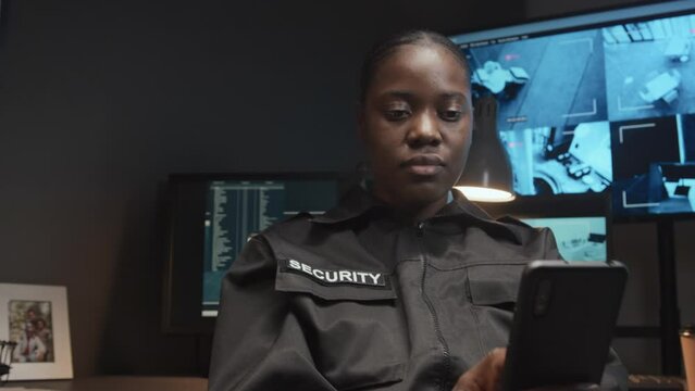 Zoom-out shot of young Black female security officer in uniform using cell phone and not looking at screens with CCTV video footage during workday in guard room