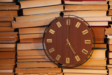 Large clock with Roman numerals on the clock face against a backdrop of stacked books shows five o'clock. End of studying, completion of education