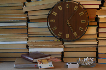 Large clock with Roman numerals on the clock face against a backdrop of stacked books, with...