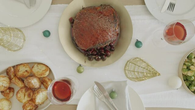 Top view 4k video of young dark skin woman putting dish with baked Christmas ham or gammon on dinner table served for Christmas eve. Festive season. Dinner table with vegetables Christmas decoration.