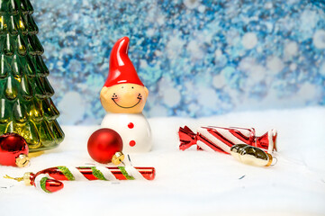 Several New Year's toys are scattered on the white snow and lie near a glass green Christmas tree and a small gnome snowman