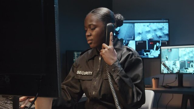 Medium shot of Black female security guard working on computer and answering work call on landline phone in surveillance room with multiple screens of CCTV footage