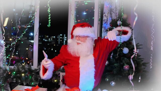 Dancing with phone in hands funny Santa Claus in red suit against background of gift boxes and Christmas tree of flickering garlands, shiny tinsel sways to rhythm of music at New Year's party.
