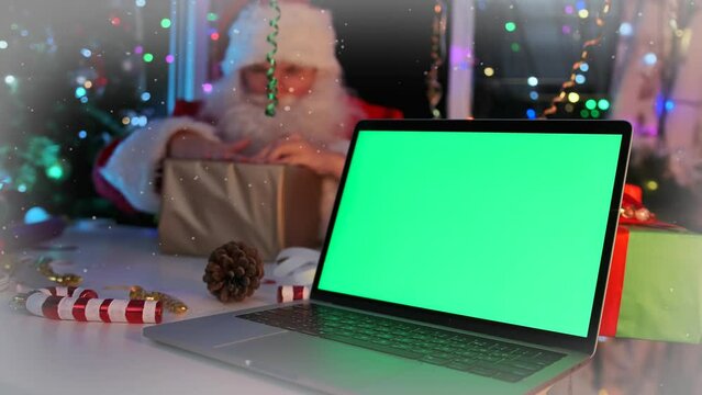 Green screen on laptop lying on table next to Santa Claus carefully wrapping gift with wrapping paper for Christmas party in room decorated with twinkling garlands of tinsel and holiday lights.