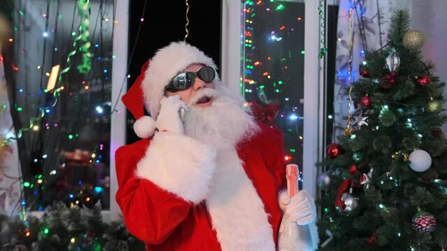 Cool modern Santa in black sunglasses with festive champagne bottle talking on smartphone while dancing at night party against the backdrop of Christmas tree and flickering garlands of festive lights.