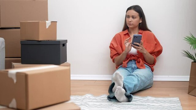Pretty woman sits with phone in hands among carton boxes telling close friends about possibility of moving to modern house with new technologies in city