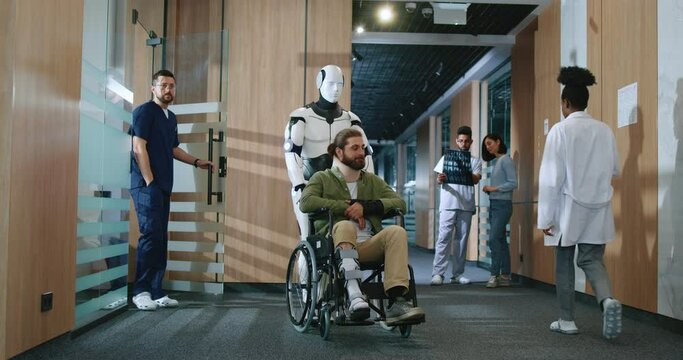 Automated robot nurse assisting young male patient with injuries and using wheelchair to navigate the hospital. Helpful robotic assistant employed in clinic. Concept for future technologies.