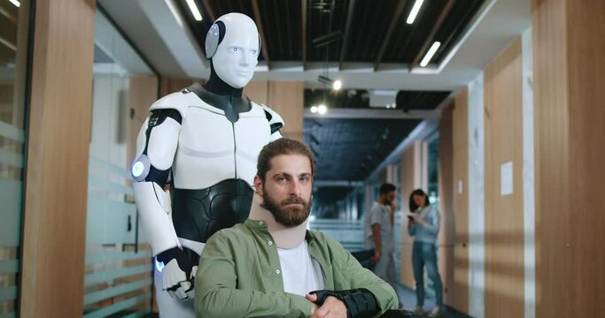 Portrait of robot assistant working at hospital helping patient standing with young handsome man sitting in wheelchair looking at camera and smiling. Future technologies concept.