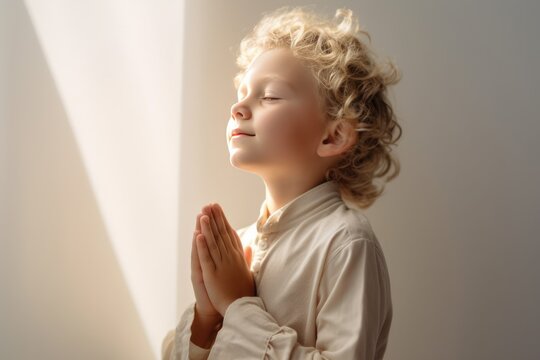 Curly little blonde boy on knees holding hands and praying in the morning, pastel neutral background. Christianity, faith, spirituality, religion, salvation, peace, faith concept. Kid praying to God