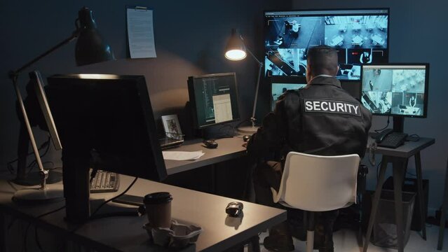 Rear view of male security officer using handheld transceiver and pointing at suspicious behavior of visitors caught on camera in surveillance room