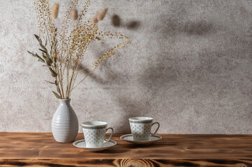 Background with bouquet of dried plants in vase, cups of coffee. Wooden shelf. Sunset light.