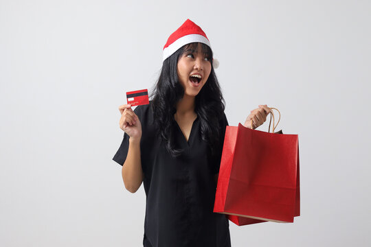 Portrait of attractive Asian woman with red Santa hat showing credit card while holding shopping bags. Cashless payment. New year and christmas concept. Isolated image on white background