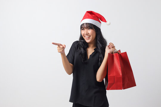 Portrait of cheerful Asian woman with red Santa hat feeling happy while pointing to the side. Excited girl holding shopping bag. New year and christmas concept. Isolated image on white background