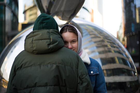 Young Girl In A Scarf On Her Head Hides On The Back Of A Guy In A Warm Jacket With A Hood And Looks From Behind His Shoulder At The Camera On The Street