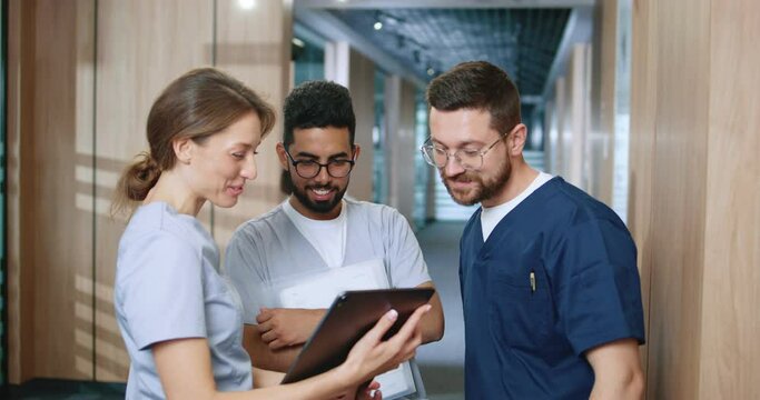 Side View Portrait Of Multicultural Group Of Young Medical Professionals Chatting During Break In Contemporary Hospital Corridor. Nurses And Doctors Using Gadgets Online.