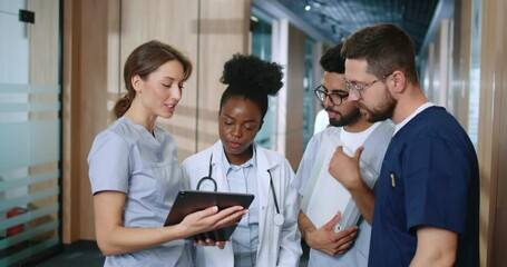 Multicultural group of youthful medical personnel communicating in modern hospital hallway during break. Nurses and doctors sharing experience and spend time using devices online. - Powered by Adobe