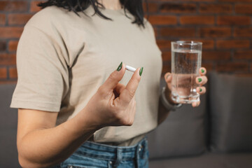 Young woman taking medicine, white pill and holds in a hand glass of water, close-up view