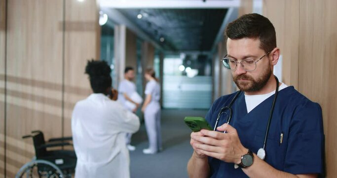 Busy and focused professional doctor in uniform using smartphone online checking social media during break in corridor of modern hospital. Successful medical worker using technologies.