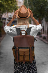 Stylish young woman in a white shirt, skirt with a craft backpack holds her hat with her hands while standing in the middle of a beautiful street, Turkey. Concept of travel, tourism.