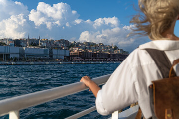 Young blonde female tourist with a craft backpack enjoys the city and the sea from a ferry boat in...