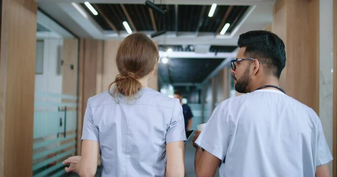 Back-view portrait of competent and talkative doctors talking and discussing their work while strolling through hospital corridors. Youthful happy male Indian nurse and Caucasian doctor at work.