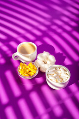 Obraz na płótnie Canvas Yellow and white colored food suplements in forms of pills is jars and a cup of coffee from above in sunlight rays on violet background.