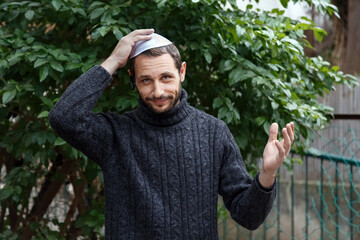 Religious Israeli caucasian man with smile face, holding a kippah, hand to his head. Handsome smiling bearded Jewish holding yarmulke  for wind over his head while looking at camera. Looks surprised - obrazy, fototapety, plakaty