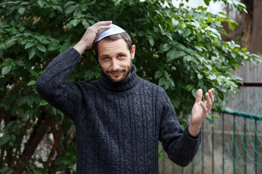 Religious Israeli caucasian man with smile face, holding a kippah, hand to his head. Handsome smiling bearded Jewish holding yarmulke  for wind over his head while looking at camera. Looks surprised