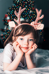 A little beautiful girl is lying on a bed with deer horns on the background of a Christmas tree.