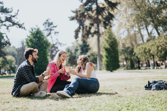 Young, carefree friends enjoy a sunny day in the park, laughing and chatting. They sit on the grass, enjoying nature and each others company. Ice cream adds to the fun in this green environment.
