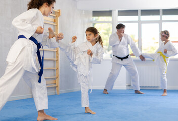 Fototapeta na wymiar Focused preteen girl in white kimono practicing karate kicking techniques during sparring with mother. Sports family activity concept..
