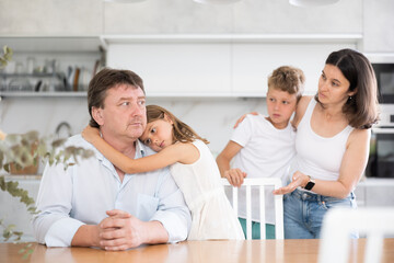 Offended father sitting at the kitchen table being hugged by little daughter, his wife and son looking at them with empathy - obrazy, fototapety, plakaty