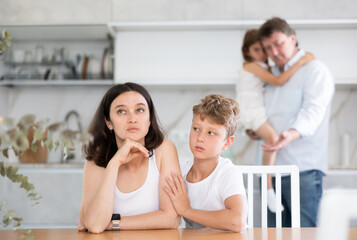 Frustrated wife sitting at kitchen table during family quarrel, with angry husband standing behind...