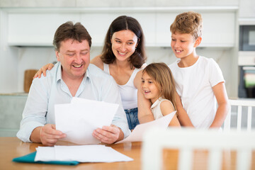 Positive husband is sitting at kitchen and reading document about career advancement at work, moneymaking financial report. Smiling wife and happy children hug successful dad