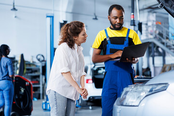 Fototapeta na wymiar Repairman at auto repair shop conducts yearly vehicle checkup, informing customer about needed engine mending. BIPOC garage worker discussing with customer after finishing car maintenance