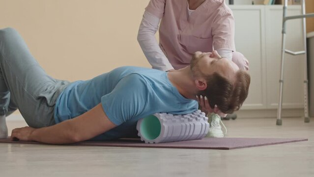 Female physiotherapist helping male patient to foam roll his back muscles during rehabilitation therapy in clinicFemale physiotherapist helping male patient to foam roll his back muscles during rehabi