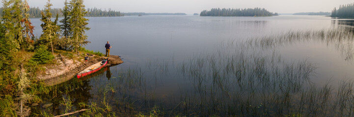 A man with a red canoe is standing on the rocky shoreline and fishing in a large lake. The northern...