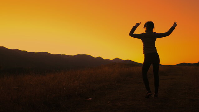 Girl dancing against bright orange sunset sky. Slim young girl silhouette having fun, moving, raise hands up, listening music in mountain field. Back view.