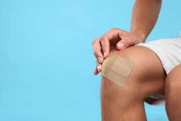 Man putting sticking plaster onto knee on light blue background, closeup. Space for text