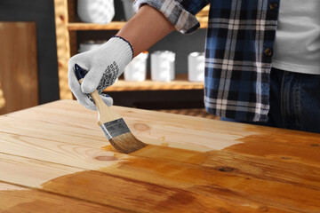 Man with brush applying wood stain onto wooden surface indoors, closeup - obrazy, fototapety, plakaty