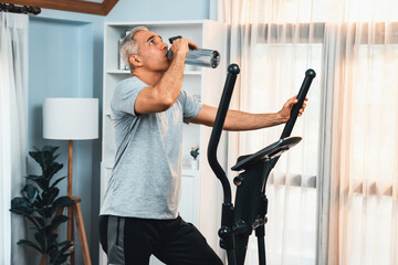 Active senior man running on elliptical running machine and drinking water at home portrait as fitness healthy lifestyle and body care after retirement for pensioner. Clout