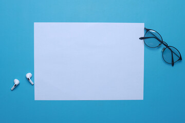 Blank white A4 paper, air pods and glasses on blue background - obrazy, fototapety, plakaty